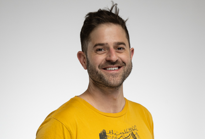 Un homme aux cheveux bruns courts et à la barbe naissante sourit à la caméra. Il porte un tee-shirt jaune orné d'un graphisme noir et se tient debout sur un fond gris clair uni.