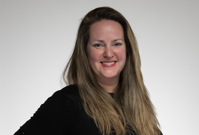 Une femme aux longs cheveux châtain clair et au haut noir sourit à l'appareil photo sur un fond gris clair uni.