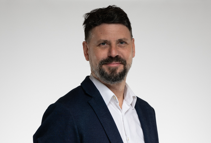 Un homme aux cheveux bruns courts et à la barbe, vêtu d'un blazer bleu marine sur une chemise blanche, se tient devant un fond clair uni et sourit légèrement à l'appareil photo.
