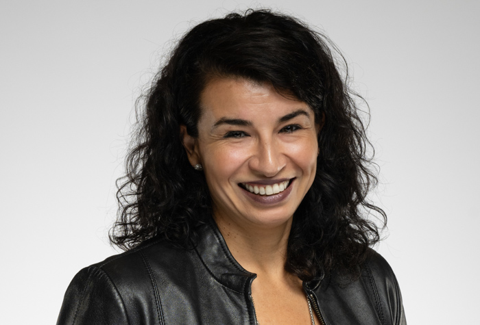 Une femme aux cheveux noirs bouclés, portant une veste en cuir noir et souriante, pose sur un fond gris clair uni.