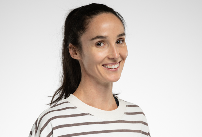 Une femme aux longs cheveux noirs attachés, vêtue d'une chemise blanche à rayures horizontales brunes, souriant et regardant légèrement sur le côté sur un fond clair uni.