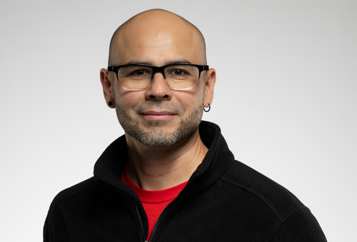 Un homme au crâne dégarni, portant des lunettes, une courte barbe et des boucles d'oreilles, est vêtu d'une veste noire sur une chemise rouge. Il sourit légèrement et pose sur un fond gris clair uni.