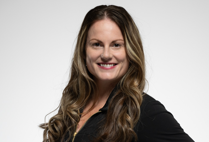 Une femme aux longs cheveux bruns ondulés avec des mèches blondes sourit à l'appareil photo. Elle porte un haut noir avec une fermeture éclair dorée et pose sur un fond clair et uni.