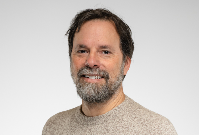 Un homme d'âge moyen aux cheveux bruns et à la barbe sourit à l'appareil photo. Il porte un pull beige et pose devant un fond gris clair uni.