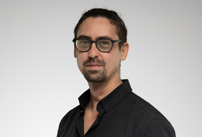 Un homme à la peau claire, aux cheveux foncés attachés, à la barbichette et aux lunettes porte une chemise foncée et regarde l'appareil photo, posant sur un fond gris clair uni.