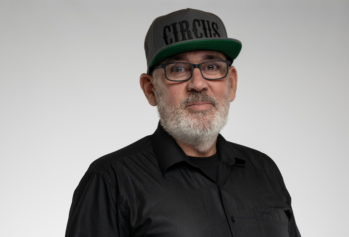 Un homme âgé avec une barbe grise et des lunettes porte une chemise noire et une casquette grise avec le mot CIRCUS imprimé dessus. Il se tient debout sur un fond gris clair uni et regarde la caméra.