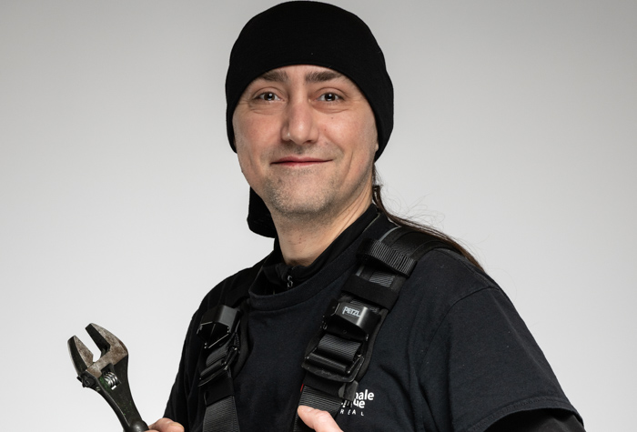 Une personne portant un bonnet noir, une chemise noire et un harnais sourit à la caméra tout en tenant une grande clé à molette sur un fond gris uni.