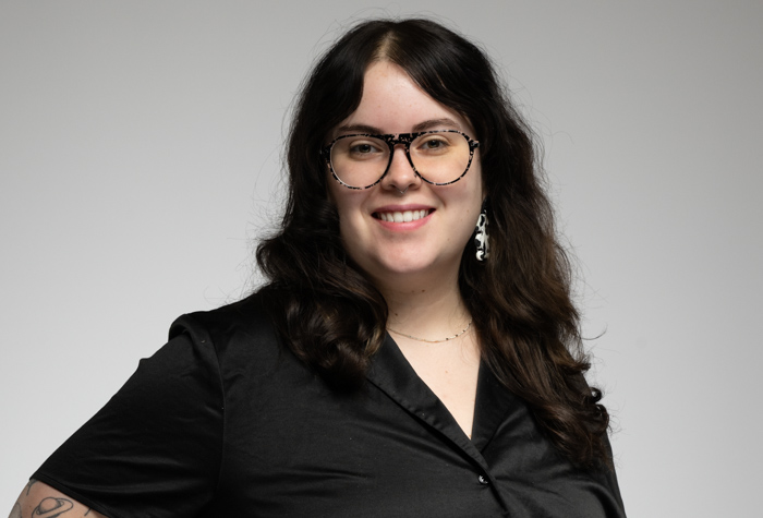 Une personne aux longs cheveux noirs ondulés, portant des lunettes, une chemise noire boutonnée et des boucles d'oreilles pendantes, sourit à l'appareil photo sur un fond gris clair uni.