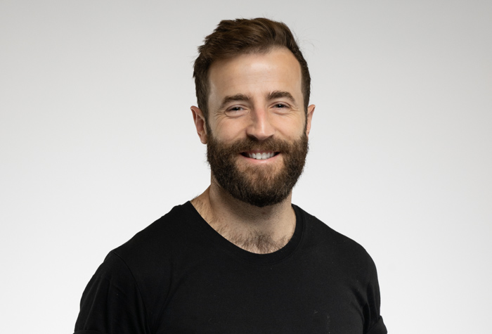 Un homme aux cheveux bruns courts et à la barbe fournie sourit à l'appareil photo. Il est vêtu d'un simple t-shirt noir et se tient debout sur un fond simple, gris clair.