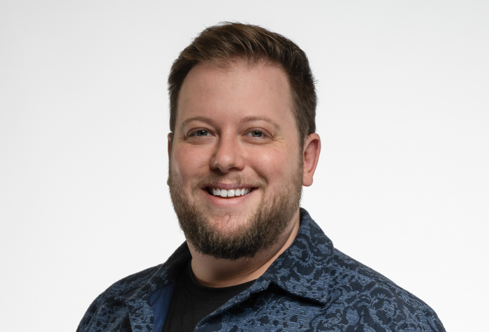 Un homme aux cheveux bruns courts et à la barbe sourit à la caméra. Il porte une chemise à motifs foncés sur un tee-shirt noir. L'arrière-plan est blanc.