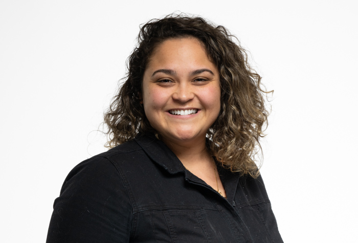 Une femme aux cheveux bruns bouclés sourit à l'appareil photo. Elle porte une chemise à col noir et pose sur un fond blanc uni.