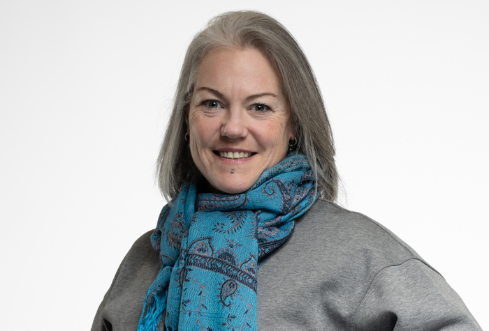 Une femme aux cheveux gris sourit à l'appareil photo. Elle porte un pull gris et une écharpe bleue à motifs, sur un fond blanc uni.