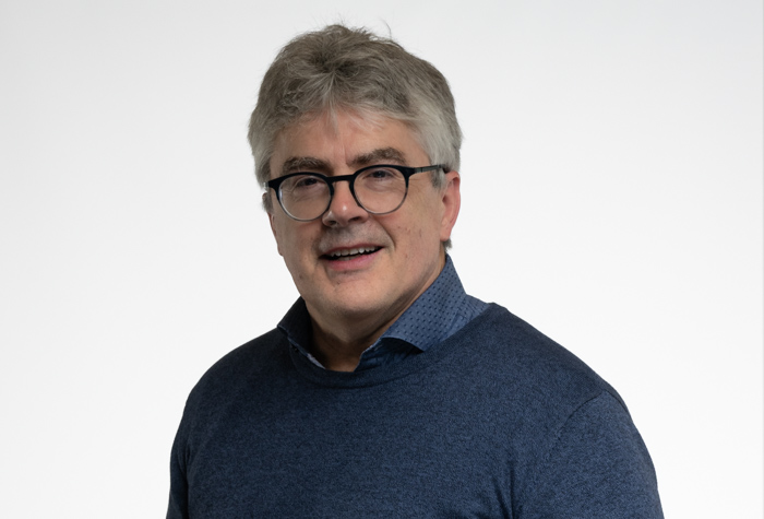 Un homme d'âge moyen aux cheveux gris et aux lunettes, portant un pull bleu sur une chemise à col, sourit à l'appareil photo sur un fond blanc uni.