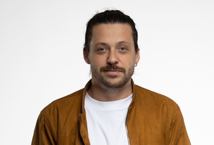 Un homme aux longs cheveux bruns attachés, à la pilosité faciale légère et à la petite boucle d'oreille, vêtu d'un t-shirt blanc et d'une veste marron, se tient sur un fond blanc uni.