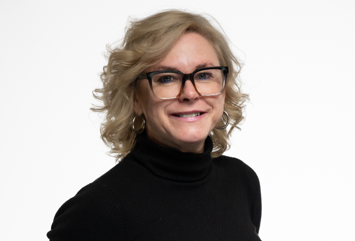 Une femme aux cheveux blonds bouclés mi-longs, portant des lunettes à monture noire, un pull à col roulé noir et des boucles d'oreilles, sourit sur un fond blanc uni.