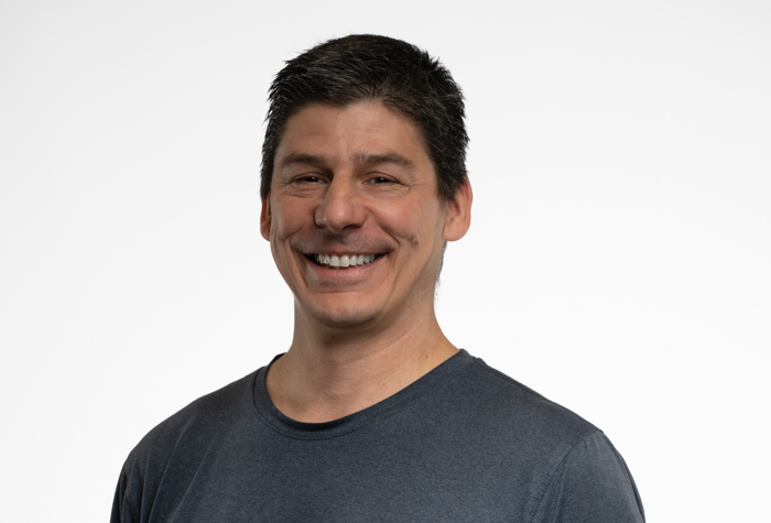 Un homme aux cheveux courts et foncés, vêtu d'un t-shirt gris foncé, affiche un large sourire sur un fond blanc uni.