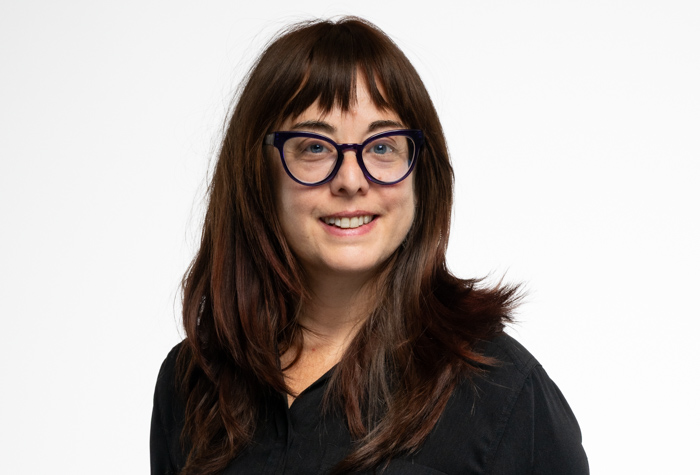 Une femme aux longs cheveux bruns et à la frange, portant des lunettes et une chemise noire, sourit à l'appareil photo sur un fond blanc uni.