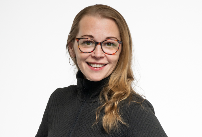 Une femme aux longs cheveux blonds, portant des lunettes et un pull noir texturé, sourit avec assurance à l'appareil photo sur un fond blanc uni.