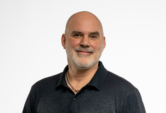 Un homme d'âge moyen au teint clair, au crâne dégarni et à la barbe grise sourit à l'appareil photo. Il porte une chemise à col sombre et un collier, sur un fond blanc uni.