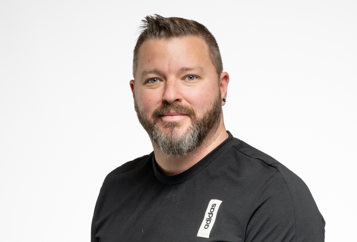 Un homme aux cheveux bruns courts et à la barbe, vêtu d'une chemise Adidas noire, sourit légèrement en posant sur un fond blanc uni.