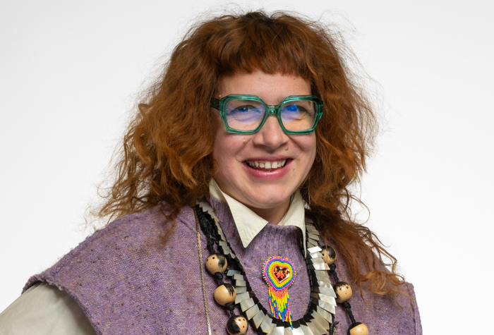 Une personne souriante aux cheveux roux bouclés, portant de grosses lunettes vertes, un haut texturé lavande, un collier de perles avec un pendentif en forme de cœur coloré, et une chemise blanche à col. L'arrière-plan est blanc.