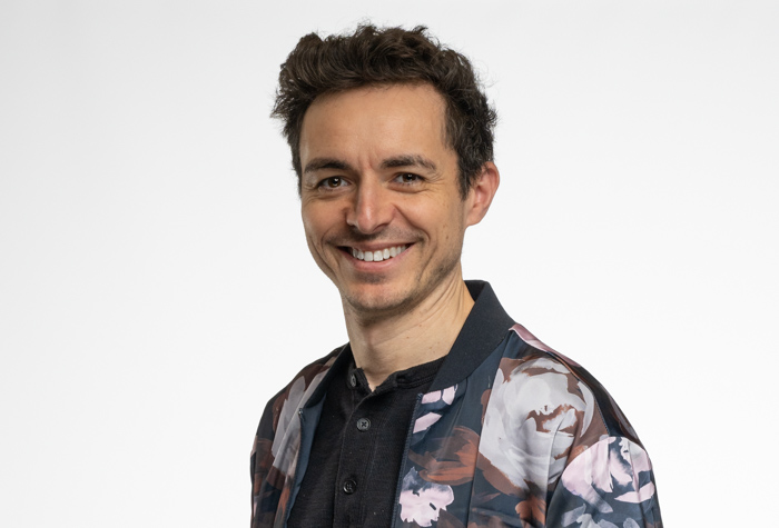 Un homme souriant aux cheveux courts et bouclés, vêtu d'une chemise à boutons foncée et d'une veste à motifs floraux, debout sur un fond blanc uni.