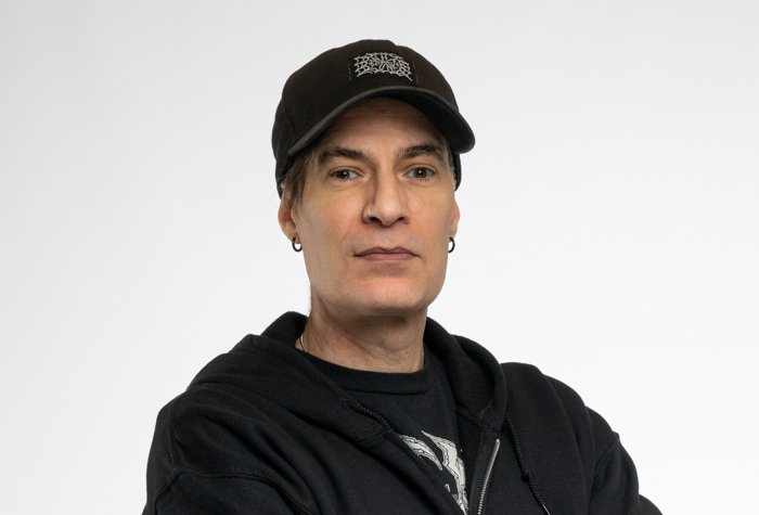 Une personne à la peau claire portant une casquette noire, un sweat à capuche noir et des boucles d'oreilles se tient sur un fond blanc uni, regardant directement l'appareil photo avec une expression neutre.