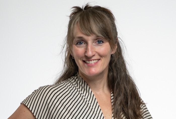 Une femme aux longs cheveux bruns et à la frange, vêtue d'un chemisier rayé noir et blanc, sourit à l'appareil photo sur un fond blanc uni.