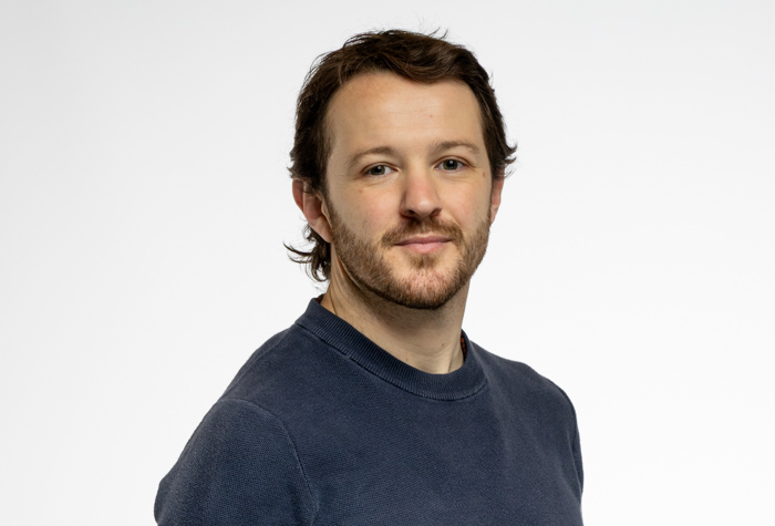 Un homme aux cheveux bruns ondulés, à la courte barbe et au pull bleu se tient sur un fond blanc uni, regardant calmement l'appareil photo.