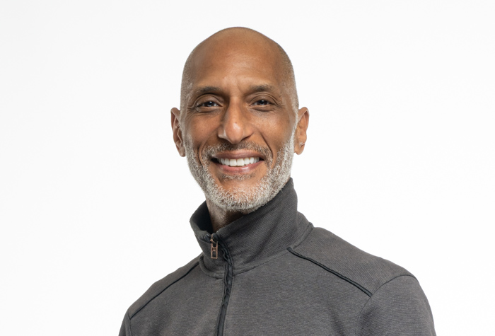 Un homme souriant d'âge moyen, au crâne dégarni et à la courte barbe grise, vêtu d'une veste sombre à fermeture éclair, se tient sur un fond blanc uni.