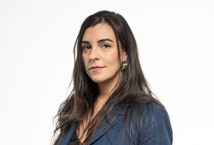 Une femme aux longs cheveux noirs, portant un blazer bleu et des boucles d'oreilles en or, se tient debout sur un fond blanc uni, regardant l'appareil photo avec assurance.