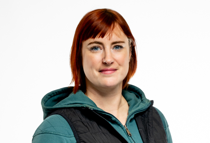 Une femme aux cheveux courts et roux, vêtue d'un sweat à capuche sarcelle et d'un gilet noir, se tient debout sur un fond blanc uni, regardant l'appareil photo avec un léger sourire.