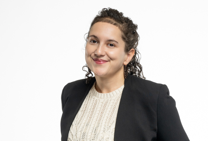 Une femme aux cheveux noirs bouclés, portant un blazer noir sur un pull en maille crème, sourit doucement devant un fond blanc uni.