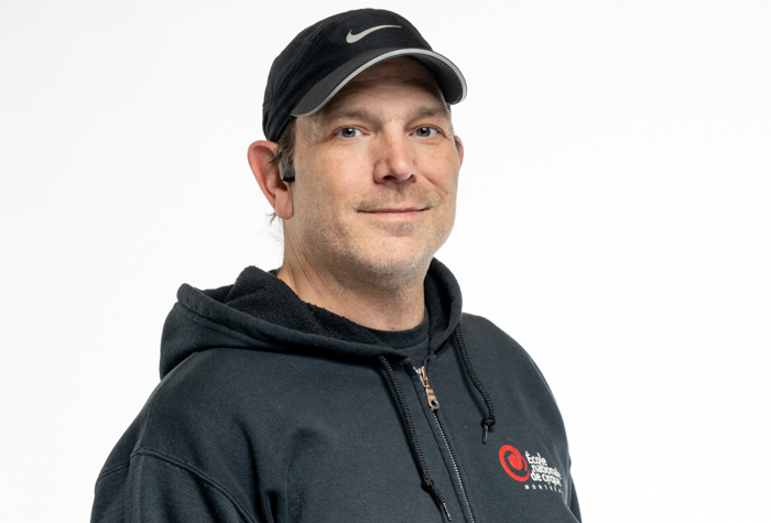 Un homme portant une casquette Nike noire et un sweat à capuche sombre avec un logo rouge et blanc, souriant à la caméra sur un fond blanc uni.