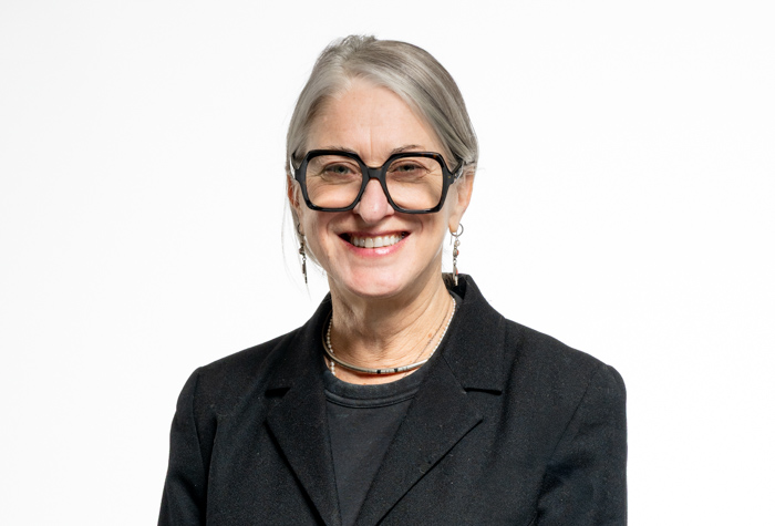 Femme souriante aux cheveux gris, portant de grandes lunettes noires, un blazer noir, une chemise sombre et des bijoux en perles, debout sur un fond blanc uni.