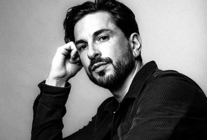 Portrait en noir et blanc d'un homme aux cheveux et à la barbe foncés, portant une veste sombre. Il appuie sa tête sur une main et regarde directement l'appareil photo avec une expression calme et réfléchie.