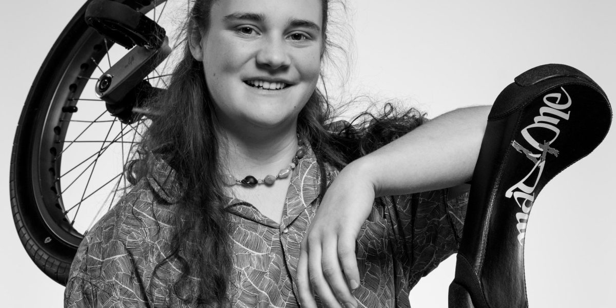 Une personne souriante aux cheveux longs, Zora Eichenberger, porte une chemise à motifs et pose avec un monocycle sur une épaule et une chaussure plate portant l'inscription Emily Wake suspendue à son bras. L'image est en noir et blanc.