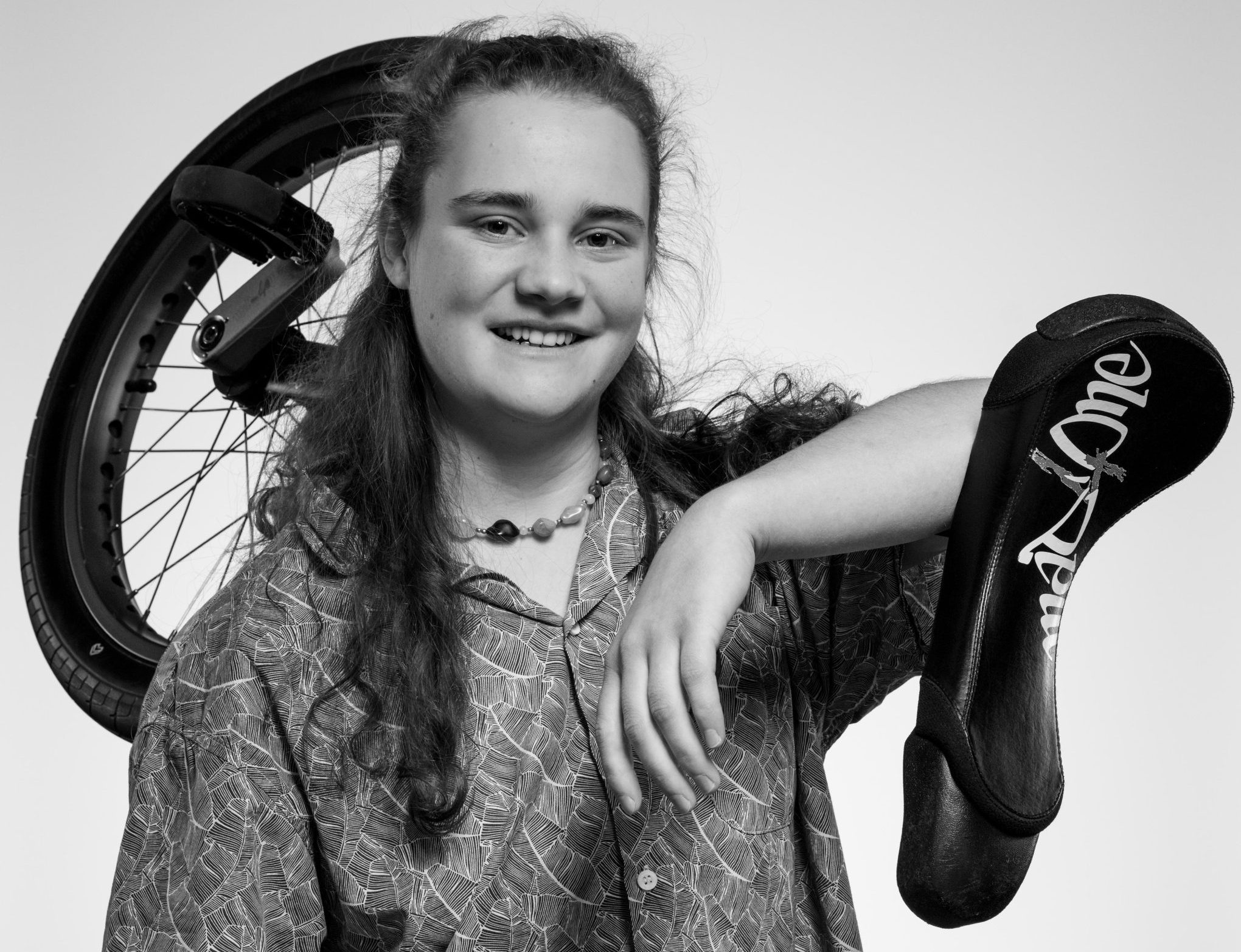 Une personne souriante aux cheveux longs, Zora Eichenberger, porte une chemise à motifs et pose avec un monocycle sur une épaule et une chaussure plate portant l'inscription Emily Wake suspendue à son bras. L'image est en noir et blanc.
