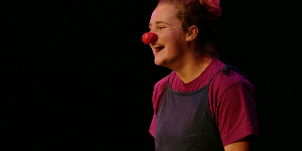 Zora Eichenberger, vêtue d'une chemise violette et d'une salopette sombre, se tient sur scène en souriant, portant un nez de clown rouge et un accessoire de coiffure rose, éclairée par les lumières de la scène sur un fond sombre.
