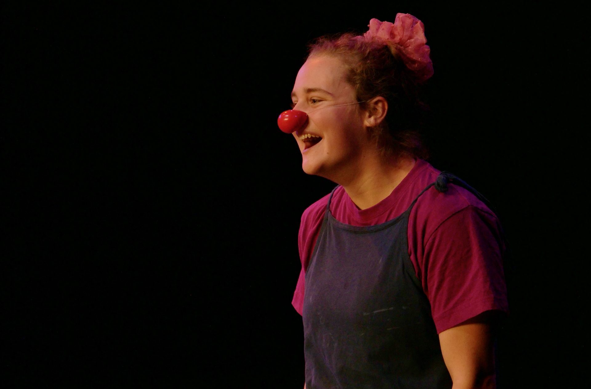 Zora Eichenberger, vêtue d'une chemise violette et d'une salopette sombre, se tient sur scène en souriant, portant un nez de clown rouge et un accessoire de coiffure rose, éclairée par les lumières de la scène sur un fond sombre.