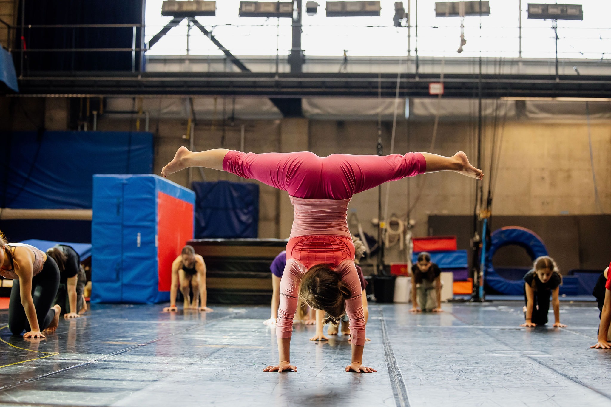 Une personne en tenue de sport rose fait un saut de mouton dans un gymnase, mettant en valeur les compétences acquises dans les cours récréatifs du SOC, tandis que d'autres s'étirent ou font des poses de yoga sur des tapis de gymnastique à l'arrière-plan.