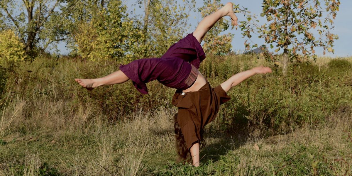 Zora Eichenberger, vêtue d'un haut marron et d'un pantalon violet, fait la roue sur l'herbe dans un champ bordé de grands arbres verts et jaunes, sous un ciel clair.