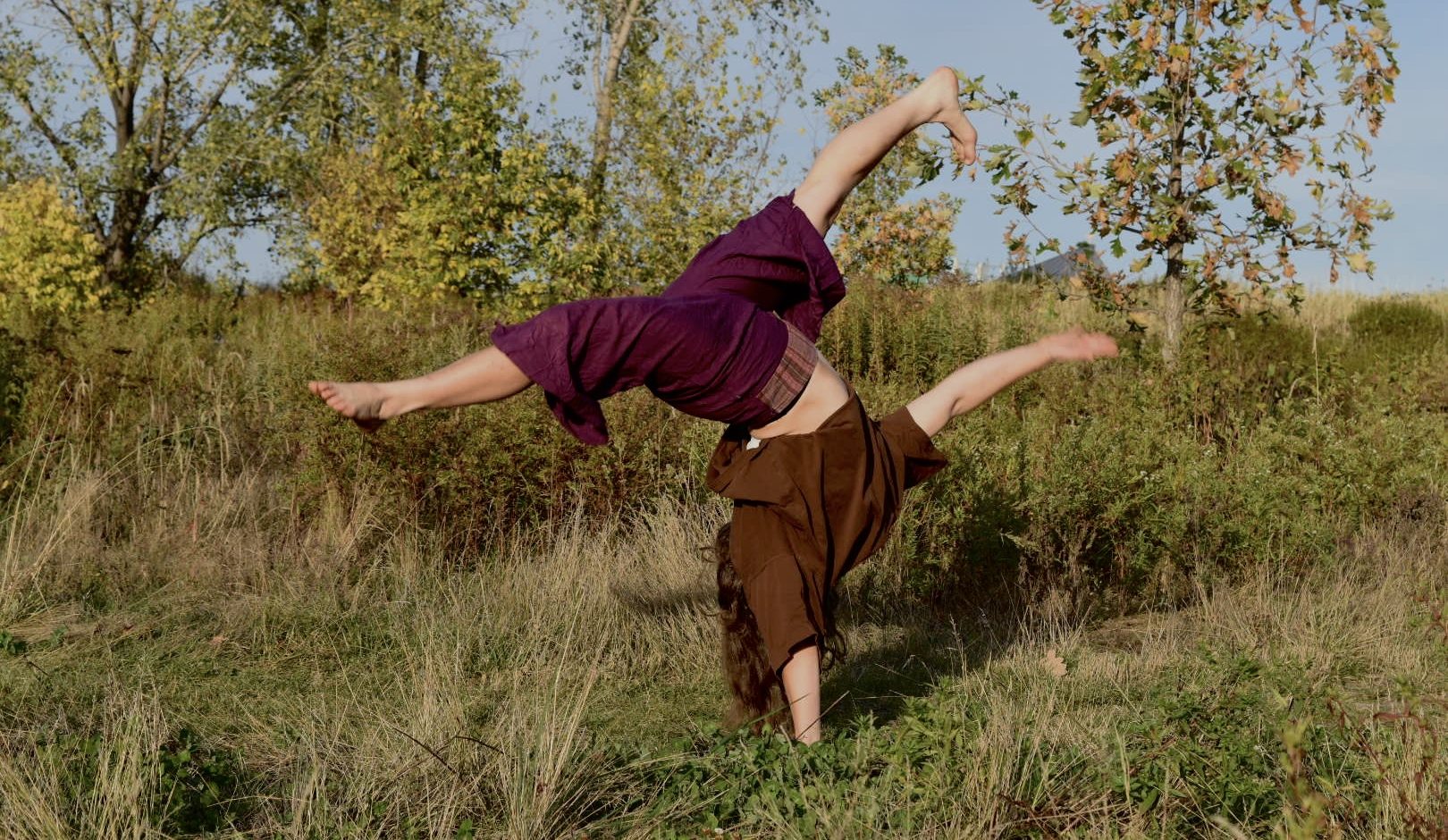 Zora Eichenberger, vêtue d'un haut marron et d'un pantalon violet, fait la roue sur l'herbe dans un champ bordé de grands arbres verts et jaunes, sous un ciel clair.