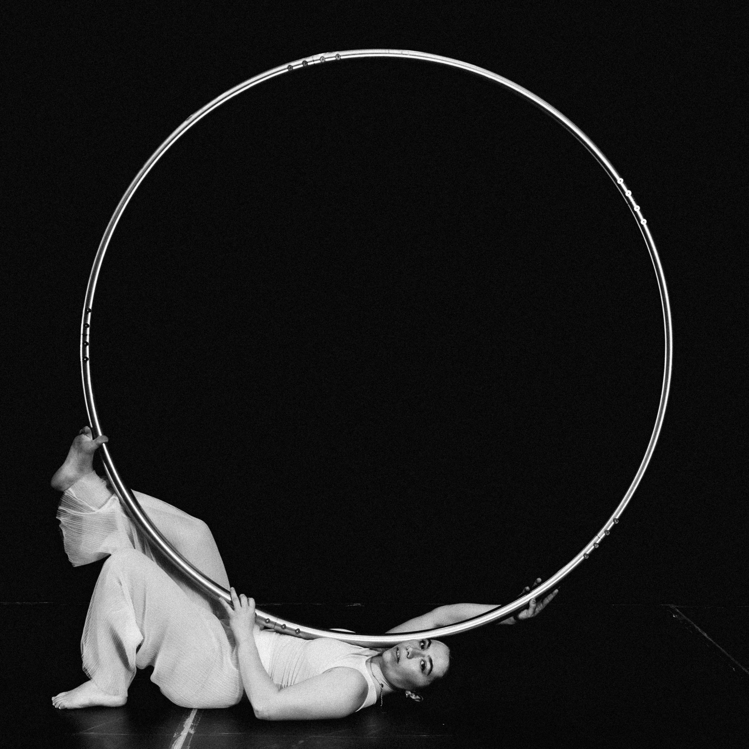Une artiste vêtue de blanc est allongée sur le sol, en équilibre et tenant un grand cerceau circulaire avec une jambe accrochée dessus, sur un fond sombre - capturant l'art gracieux de Léonie Salcines.