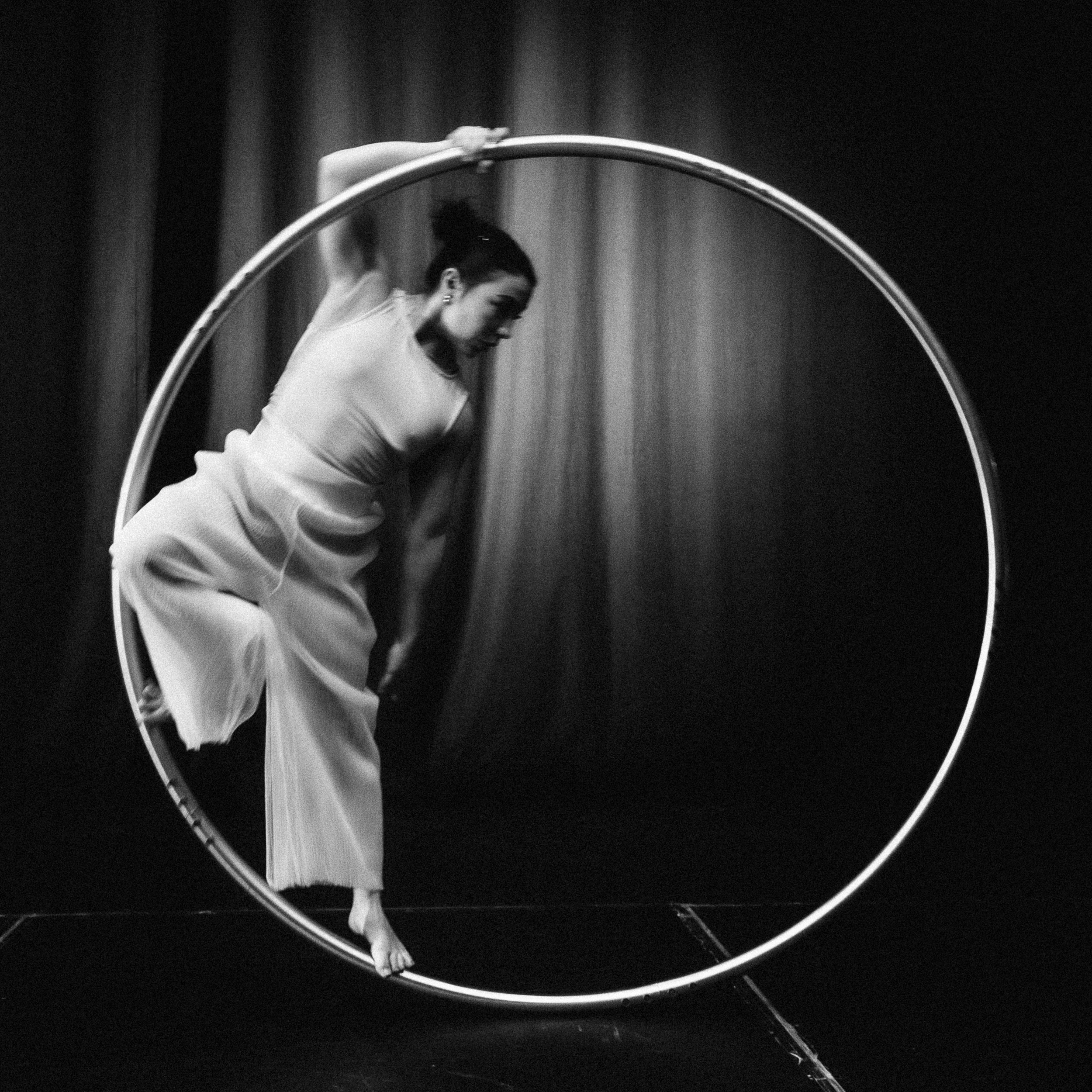Une personne vêtue d'une tenue fluide se balance gracieusement à l'intérieur d'une grande roue de cirque, connue sous le nom de roue Cyr, sur une scène sombre avec en toile de fond un rideau légèrement éclairé. Cette image saisissante en noir et blanc met en scène la talentueuse artiste Léonie Salcines.