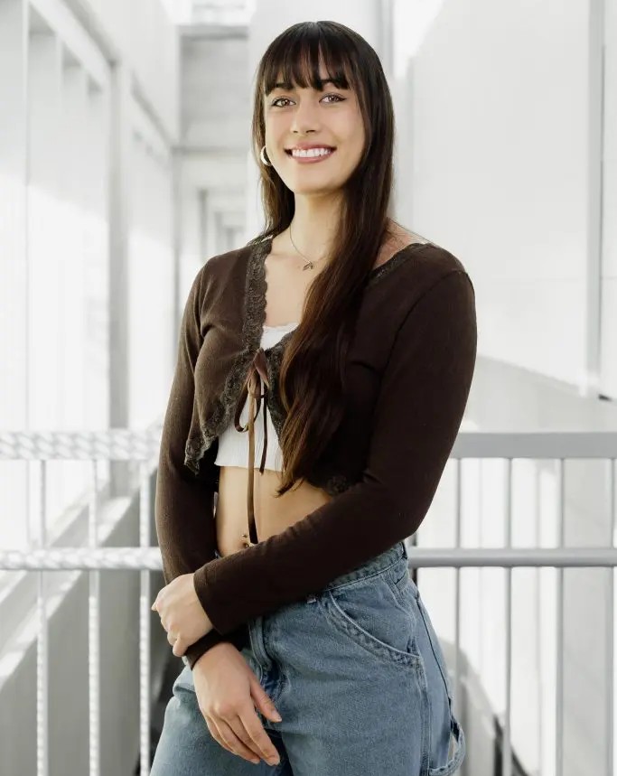Moehana Vanaa, une jeune femme aux longs cheveux châtains et à la frange, vêtue d'un cardigan court marron par-dessus un haut blanc et un jean bleu, se tient debout, souriante, dans un couloir lumineux et moderne baigné de lumière naturelle.