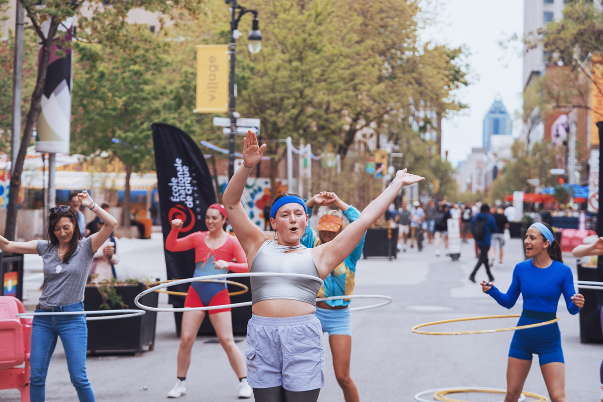 Un groupe de personnes faisant du hula hoop en plein air dans une rue de la ville, souriant et levant les bras dans un préambule animé. Des arbres et des bannières bordent la rue, tandis que d'autres personnes marchent à l'arrière-plan, ajoutant à l'atmosphère énergique.