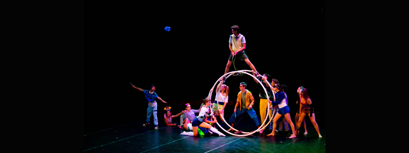Un groupe d'artistes forme une figure acrobatique sur scène : l'un d'entre eux se tient debout au sommet d'un grand cerceau lumineux, tandis que les autres le soutiennent, maintiennent l'équilibre ou interagissent avec énergie autour et à l'intérieur du cerceau, sur fond sombre.