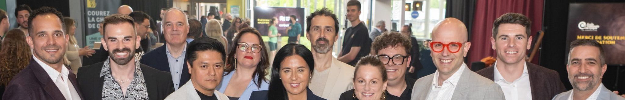 Un groupe de douze personnes, élégamment vêtues en tenue professionnelle ou semi-formelle, sourient pour une photo lors de la Soirée-bénéfice 2026, au cours de laquelle 150 000 dollars ont été récoltés pour soutenir la prochaine génération à l’École nationale de cirque. Des affiches et une enseigne de bistrot ajoutent du charme à cet espace événementiel moderne.