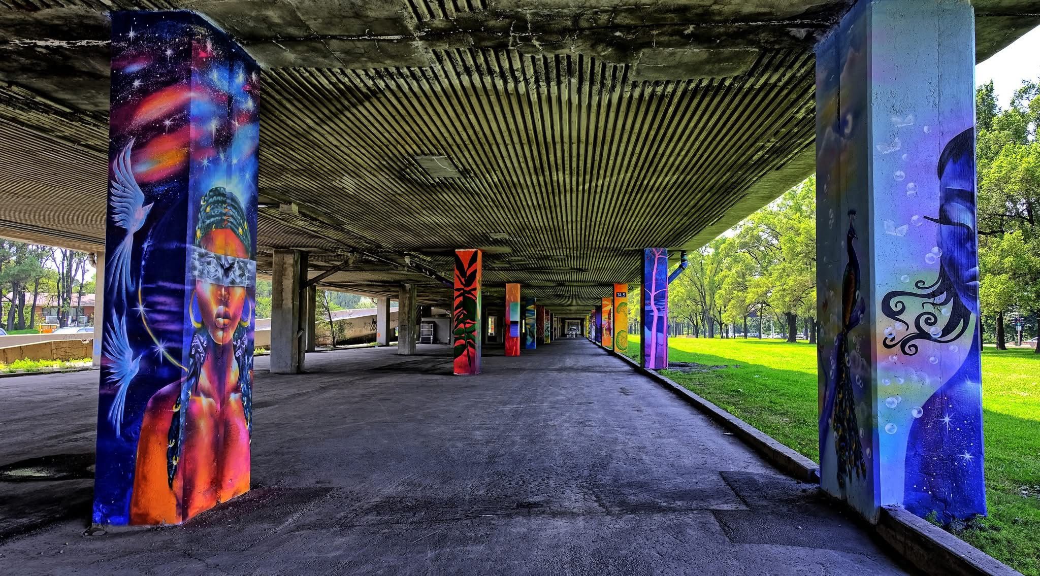 Les piliers en béton situés sous une autoroute sont recouverts de fresques murales aux couleurs vives sur le thème de l'univers, représentant notamment des visages abstraits et des motifs célestes. Des arbres et de l'herbe entourent la structure, créant une atmosphère inspirante qui rappelle celle du Quartier des arts du cirque, qui dévoile sa vision de développement pour le secteur Saint-Michel.
